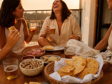 Amigas rindo em volta da mesa de petiscos numa varanda, no fim de tarde de sábado
