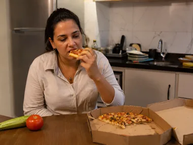Mulher comendo pizza de delivery na mesa da cozinha, com os legumes que ela comprou intactos ao lado